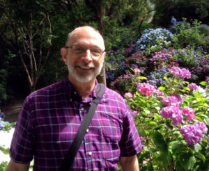 Fr. Nick in a garden in Wellington, New Zealand, wearing a short-sleeved purple shirt with purple rhododendrons in the background.